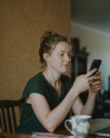 A woman at a wooden desk reading her phone, with a mug, a notebook and a laptop on the table and a plain ochre wall behind her.