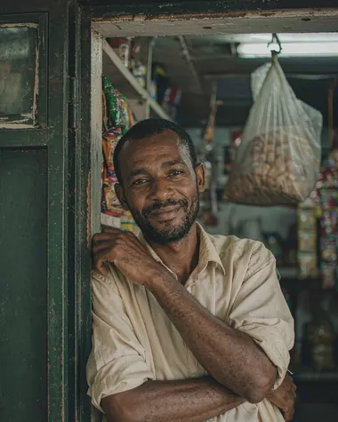 A man in a pale shirt leaning in the doorway of his provisions store with his arms folded, glancing towards the camera, shelves of goods behind him.