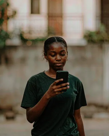 A woman in a dark green t-shirt walking along a residential street, looking down at the phone in her hand.